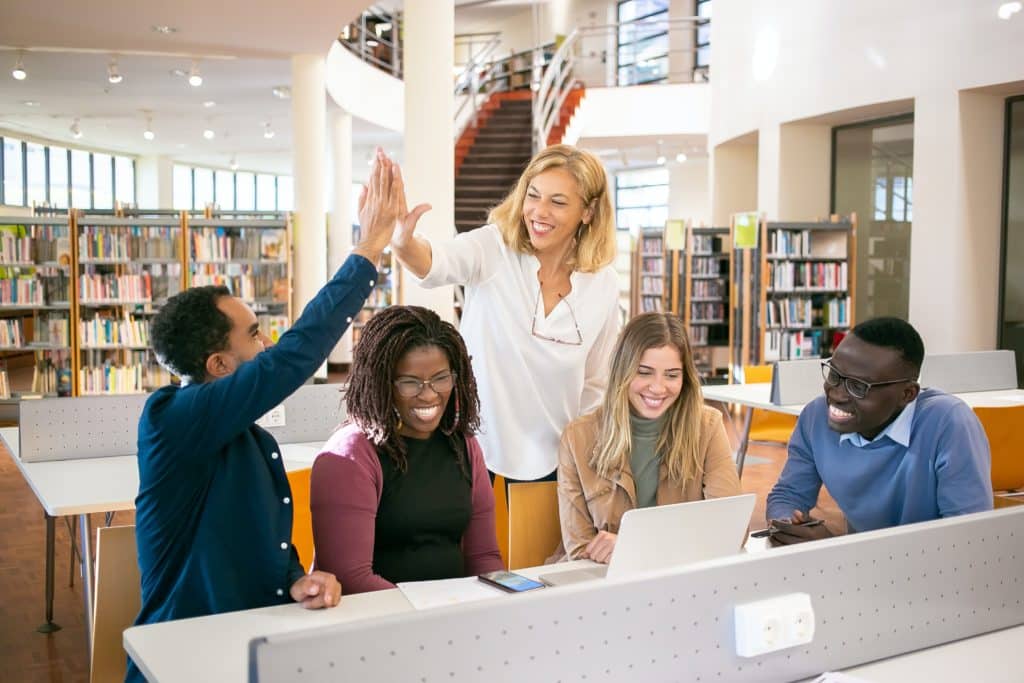 A picture of a team sitting around a computer in a library while two professionals are high-fiving. This article is about the best project portfolio management solutions also known as PPM software.